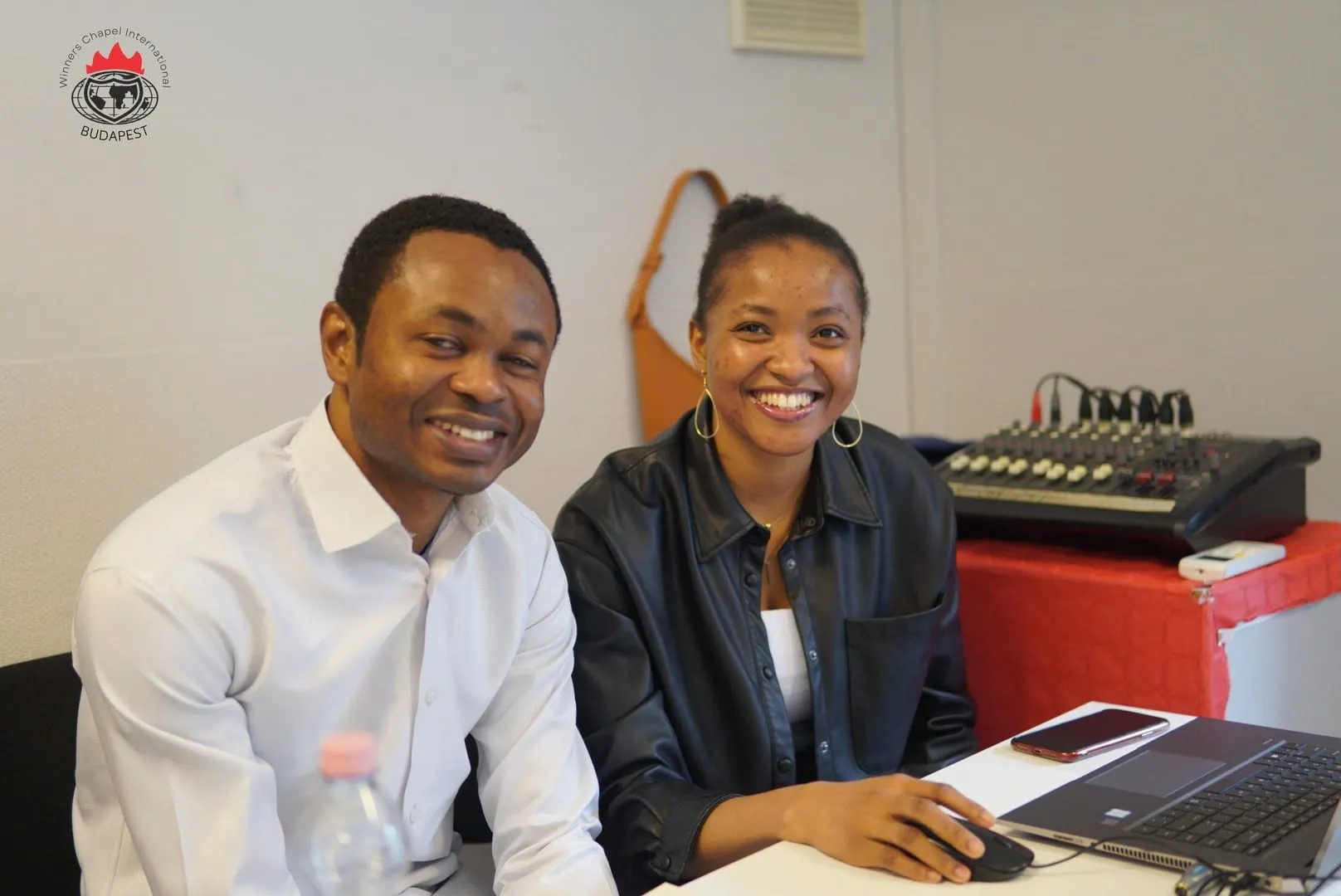 Two volunteers smiling at the media desk beside a sound mixer and a laptop