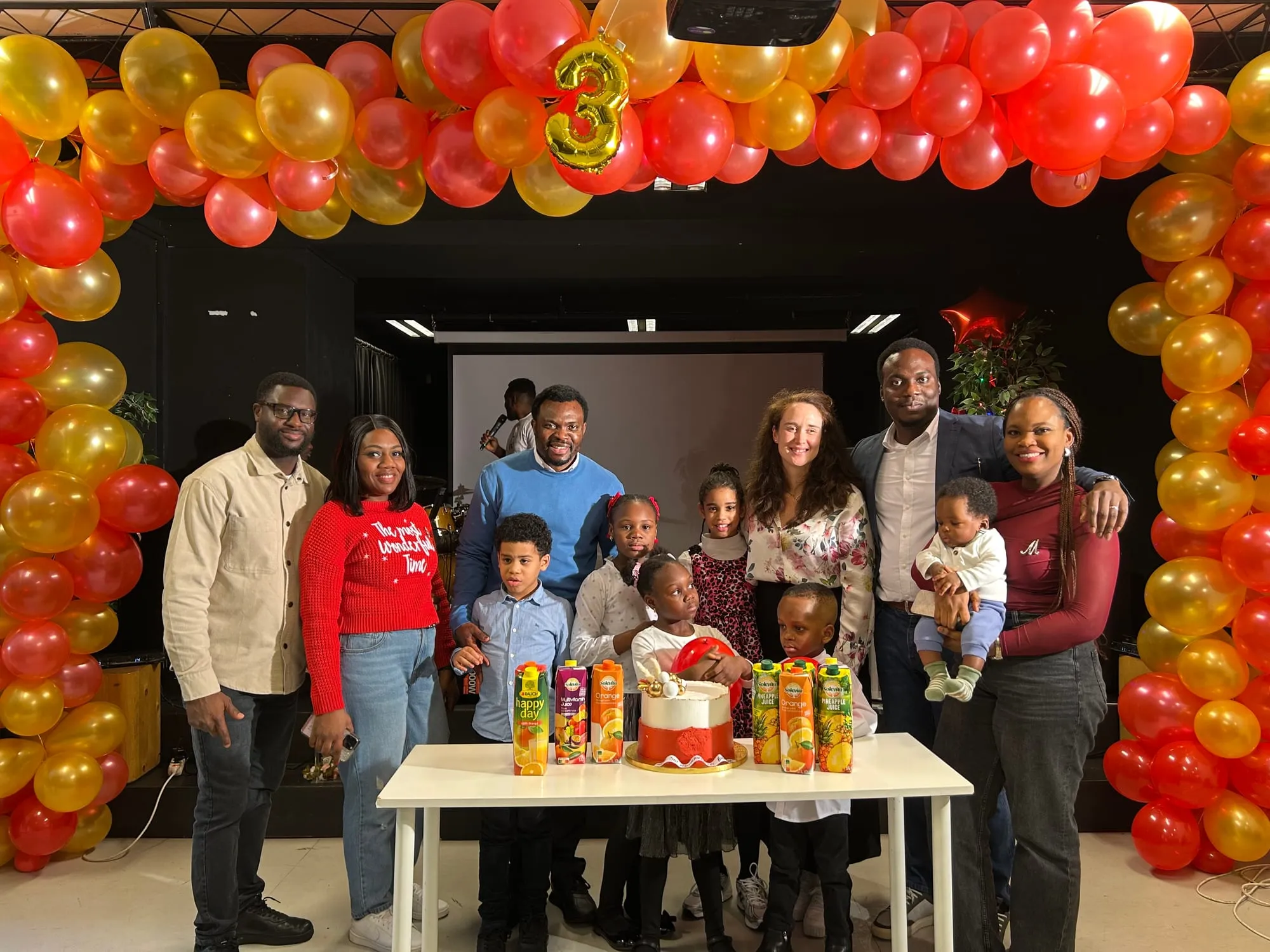 Children and their teachers standing behind a cake under a balloon arch