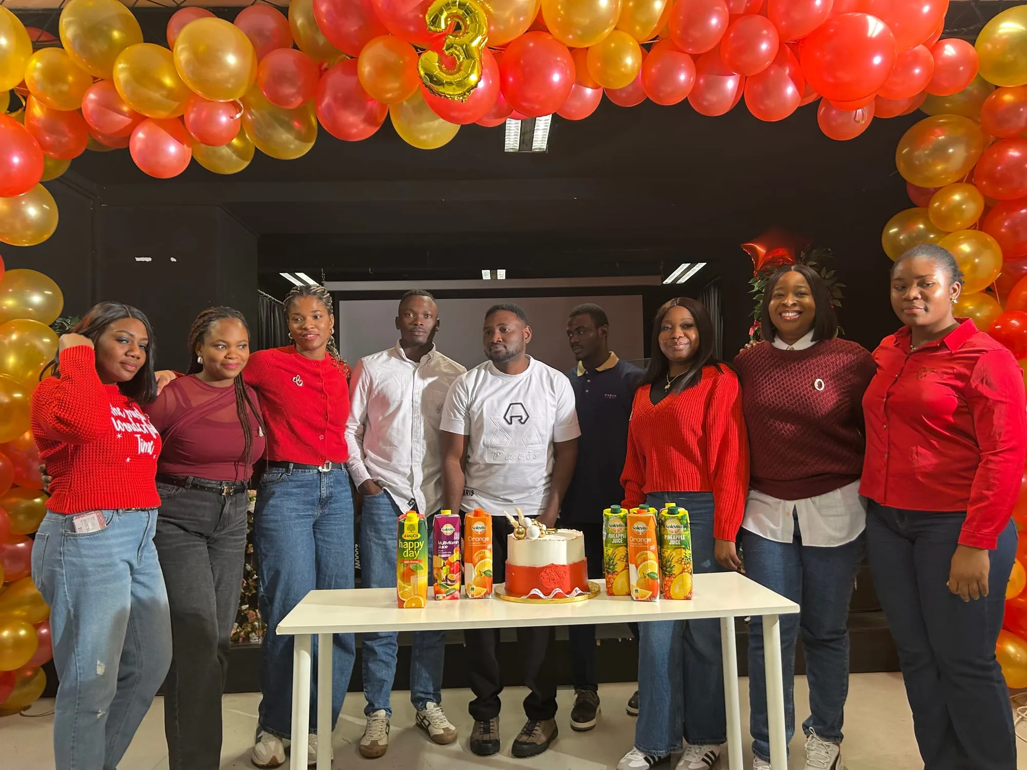 Nine young adults standing together behind a cake under a balloon arch